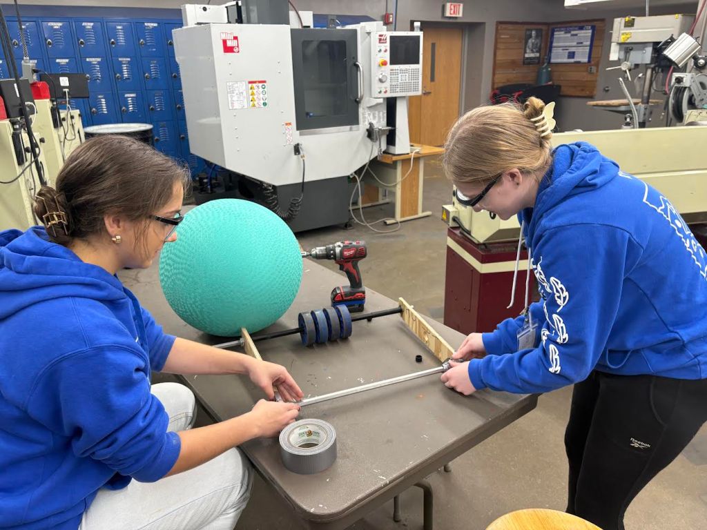 A workshop scene with Rainee R. Kali M. collaborating on their first prototype. Both are wearing blue Chicken Bot Pie hoodies and are measuring a metal rod with a tape measure. The workbench in front of them holds various tools and materials, including a roll of duct tape, a large teal kickball (which is the algae), a power drill, and wooden planks. 
