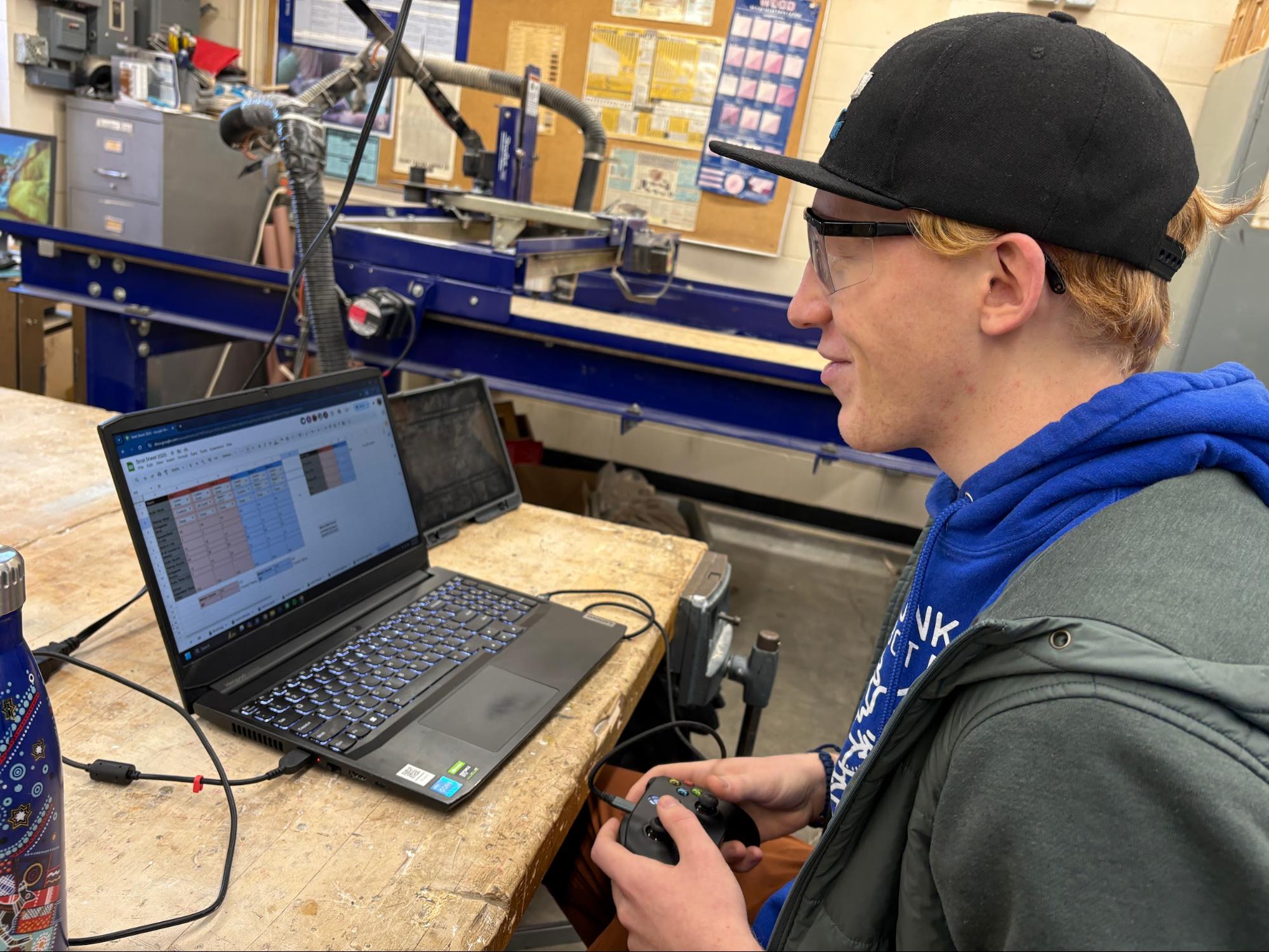 Evan G, wearing safety glasses, a blue team hoodie, and a gray hoodie over it, is seated at a wooden table in a workshop environment, using a laptop. The laptop screen displays a spreadsheet with various columns and rows of data. Evan is holding a game controller in both hands. 