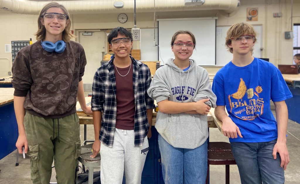 4 members of the build team in the wood shop wearing safety glasses smiling for the camera 