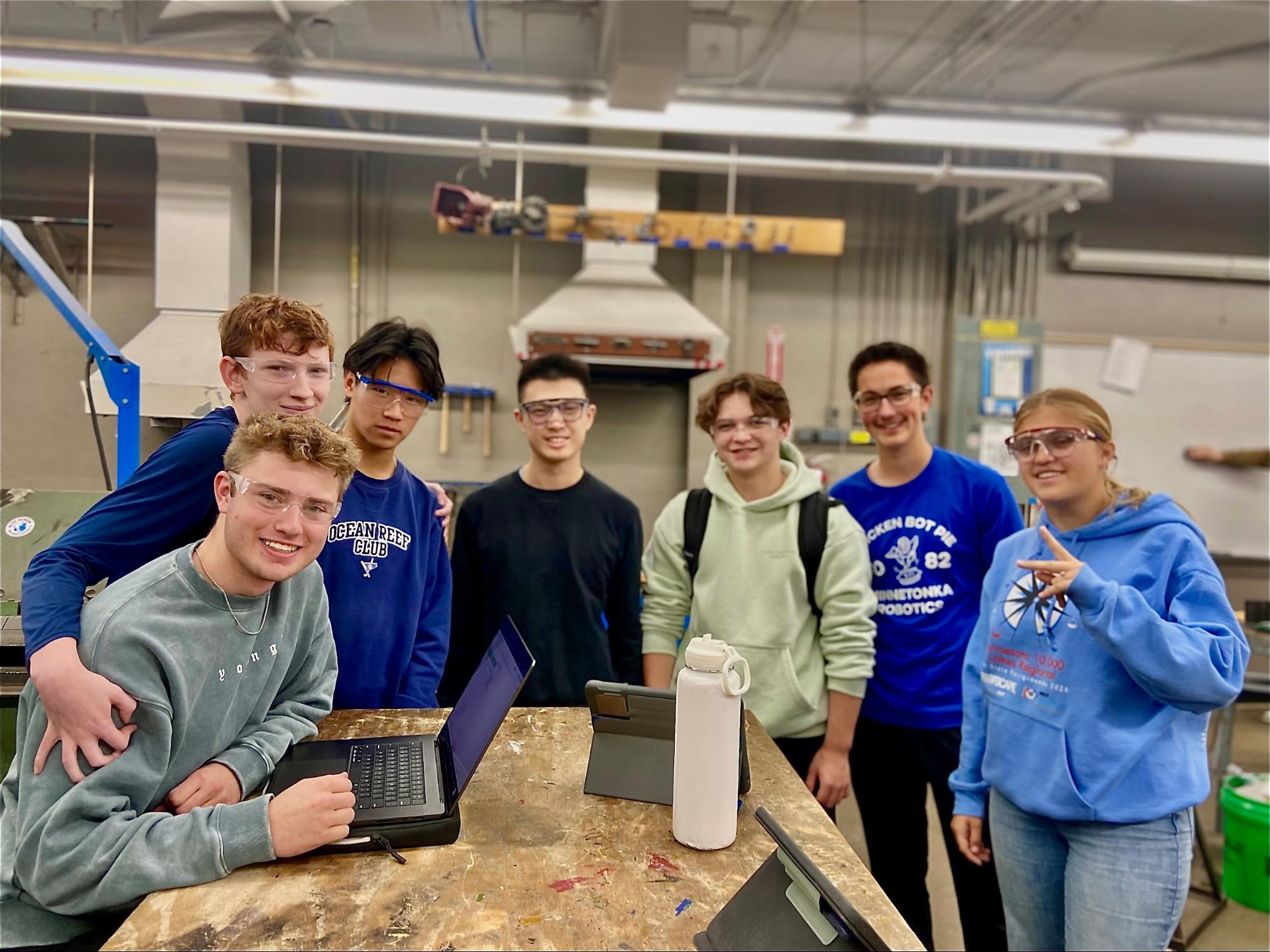 The strategy team huddled up taking a group photo in the metal shop. They look excited to work with their devices which are in front of them on a table.